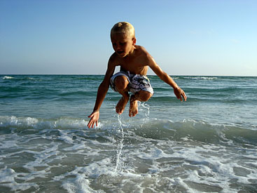 Vert: Boy jumping high on ocean shore